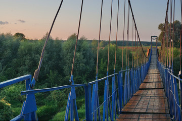 Hanging (suspension) bridge across a narrow river. Blue iron fences and old wooden bottom. Summer. Green trees and meadows. Clear blue sky. Evening. Middle Russia. Kaluga region