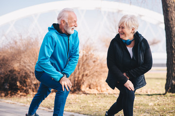 An elderly couple stretching their knees at the park. They are heartfully smiling. The woman is wearing blue headphones around her neck.