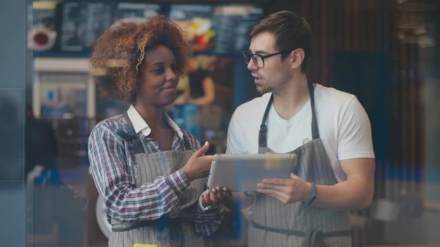 Smiling diverse waiter and waitress using digital tablet standing in cafeteria