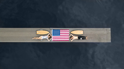 Young couple of surfers lies on a wooden pier near the beach among the calm ocean waiting for the wave. American flag and sports fitness people. Surfboards girl and guy. Top View.