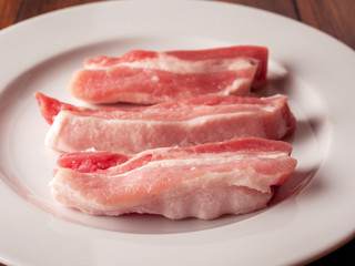 Uncooked raw pork belly slices on a white plate and wooden table.