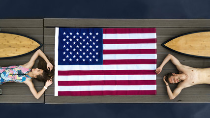 Young couple of surfers lies on a wooden pier near the beach among the calm ocean waiting for the wave. American flag and sports fitness people. Surfboards girl and guy. Top View.