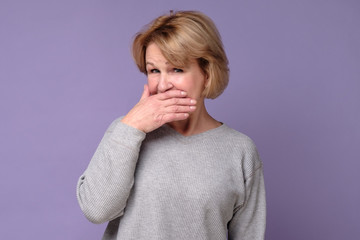 Senior older woman with puzzled look covering her mouth trying to keep secret. Studio shot