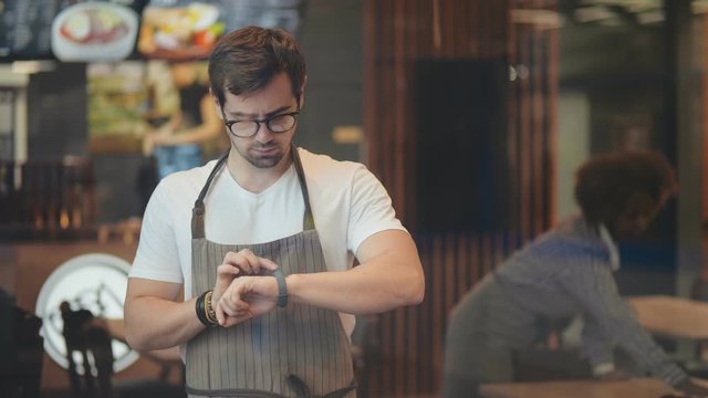 Young cafe owner using smartwatch to pay online for supplies standing in cafe hall.