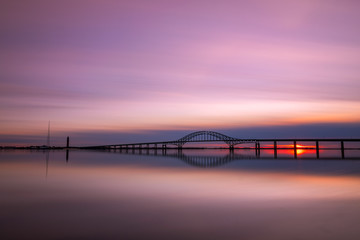 Fototapeta na wymiar Steel tied arch bridge spanning a bay with crystal clear reflections in the water at sunset. Fire Island Inlet Bridge, part of the Robert Moses Causeway on Long Island New York. 