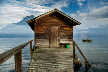 Wooden boat house with wooden footbridge on Traunsee lake with Traunstein peak on background covered with low clouds, located in Austria, Alpine region.