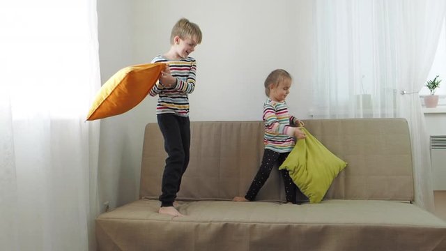Brother and baby sister fighting by pillows on the sofa, slow motion