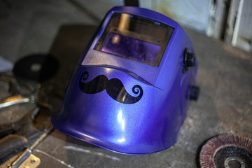 Mask for protection against bright welding. Mustache glued on a mask. An adhesive film was used to create a mustache sticker. Work in the workshop. The husband loves manual labor.