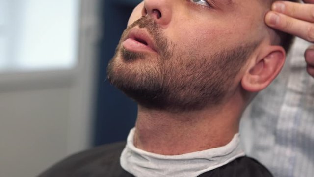 Barber shaves the client's beard on a chair. Beard haircut. Barber to shave a beard with an electric razor. Grooming of real man. Side view of young bearded man getting beard haircut at hairdresser