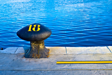 Number 18, eighteen, stencil yellow digit painted on a big commercial harbor bollard at the edge of...