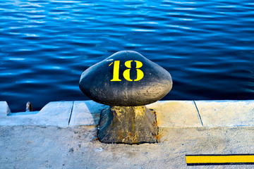 Number 18, eighteen, stencil yellow digit painted on a big commercial harbor bollard at the edge of...