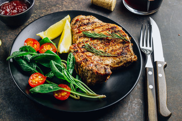 Roasted pork steak on black plate over dark background. With lemon, Cranberry Sauce and wine.