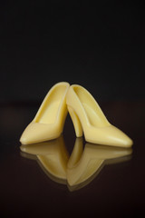 Tiny yellow high-heeled doll shoes are reflected on a black background and surface.