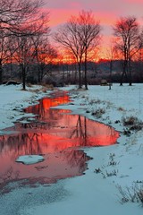 Peaceful, sunrise photo of a Midwest winter countryside scene with a creek running through a field, snow on the ground, and trees in the background