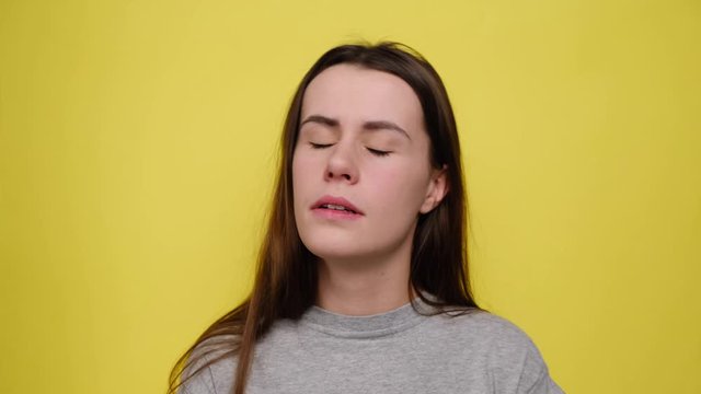 Unhappy depressed young woman showing finger gun gesture at her temple, dressed in grey t-shirt, isolated over yellow background. Negative human emotions and feelings concept