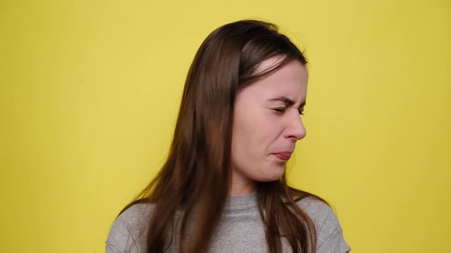 Unhappy woman frowns face, has disgusting expression, shows tongue, irritated with somebody, dressed in grey t-shirt, isolated over yellow background. People and negative facial expressions