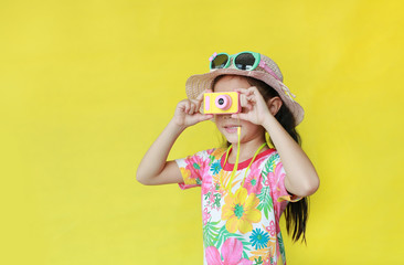 Asian little kid girl in floral pattern summer dress and hat with sunglasses taking photo by toy camera isolated over yellow background. Holiday and summer fashion concept.