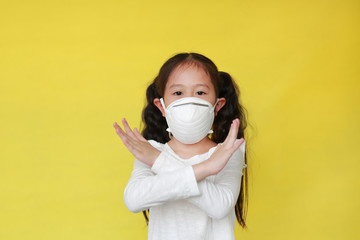 Asian little child girl wearing a protection mask with making X sign with her arms against air pollution isolated on yellow background. Kid with health care concept.