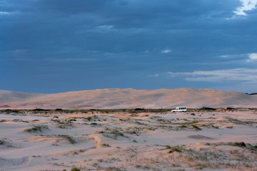 Wide boundless flat desert and a point of a white car driving along horizontal stripe of a road far away. Stockton Sand Dunes near the coast, Worimi Regional Park, Anna Bay, Australia