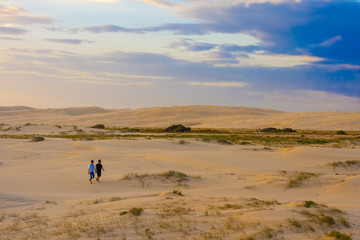 Two people in T-shirts and shorts are walking in the desert. Stockton Sand Dunes near the coast, Worimi Regional Park, Anna Bay, Australia