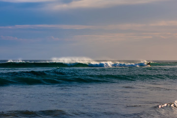 Low wave in the ocean, the spray from her beautifully sprayed in the air. The silhouette of a surfer in the distance. Worimi Regional Park, Anna Bay, Australia