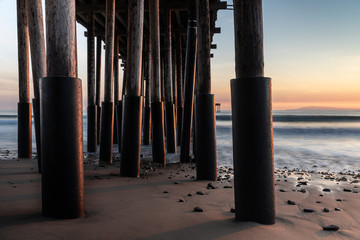 Closeup of pillars, Ventura Pier, Ventura, California at sunset. sand and rocks in foreground; silky ocean, colored sky beyond. 