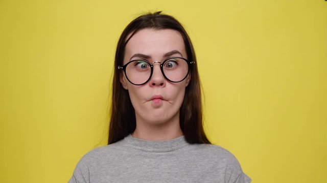 Crazy comic female in glasses makes funny face, crosses eyes, plays fool, doesnt want to be responsible, wears grey t-shirt and stands over yellow background. Facial expressions.