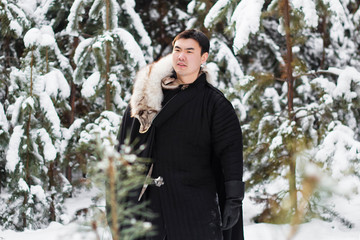 A warrior wearing black quilted clothes in a black cloak with a white fur collar and a long sword in his hands is walking along deep snow against the backdrop of a pine forest.