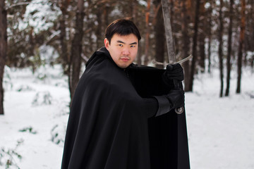 Fototapeta premium portrait of a warrior in black quilted clothes, leather gloves, a cloak with a hood and a sword in his hands against the backdrop of a winter forest