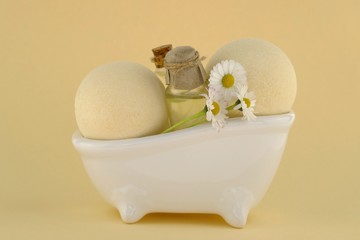 Bath bombs with chamomile extract.Organic natural body cosmetics. Two bath bomb balls and daisy flowers in a decorative white bath on a light yellow background.Cosmetics for bath.