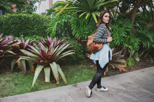 Urban Woman Walking With Backpack Bag On The Sidewalk With Garden In The Background