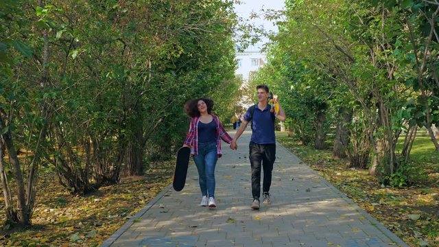 Content young male with skateboard holding hands with optimistic plump girlfriend walking in autumnal park looking away