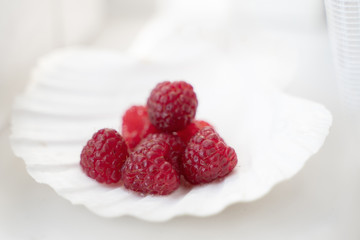 a handful of raspberries lies in a mother of pearl plate in the form of a shell on white table on white background