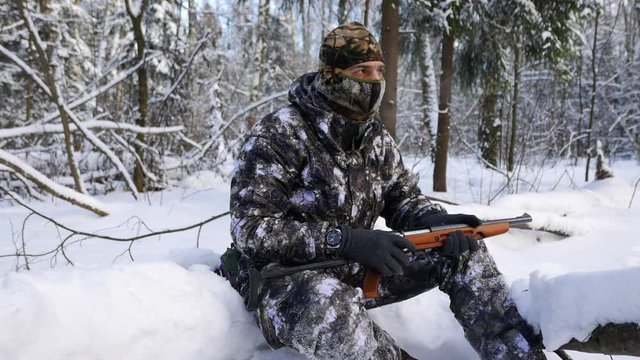 A hunter in a camouflage suit sits on a fallen tree with a gun in his hands in a snowy forest. Frosty winter.