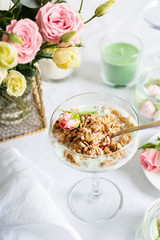 Light granola with Greek yogurt in a glass goblet, perfectly preserved on a white tablecloth amid flowers and sweets.