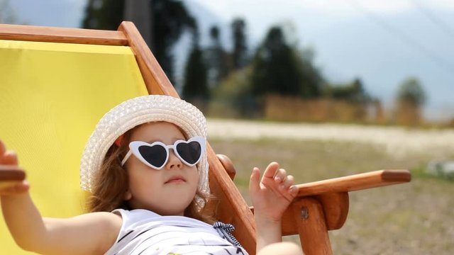 Tourist baby girl is sitting on a deck chair and waving hand. Baby girl with sunglasses and a hat eats cand y and relaxes on a journey.