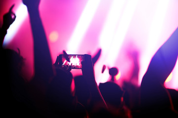 Naklejka premium Hand with a phone records live music festival. People taking photographs with smart phone during a public music concert. Crowd raising their hands, dancing and enjoying great the concert.