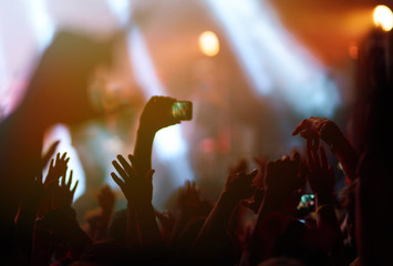 Hand with a phone records live music festival. People taking photographs with smart phone during a public music concert. Crowd raising their hands, dancing and enjoying great the concert.