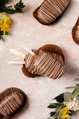 Chocolate pop cake in the form of popsicle with pink and yellow spring flowers on a marble background. Top View Image.