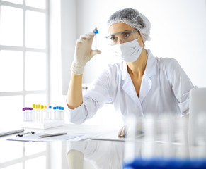 Professional female scientist in protective eyeglasses researching tube with reagents in...