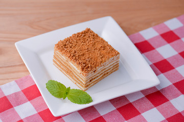 A slice of layered vanilla honey cake on a white plate with a mint leaf on wooden table with a plaid table cloth