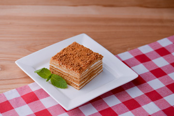 A slice of layered vanilla honey cake on a white plate with a mint leaf on wooden table with a plaid table cloth