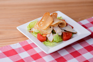Traditional classic Caesar salad with chicken served in a white plate over light wooden table with plaid tablecloth.