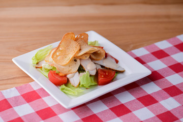 Traditional classic Caesar salad with chicken served in a white plate over light wooden table with plaid tablecloth.