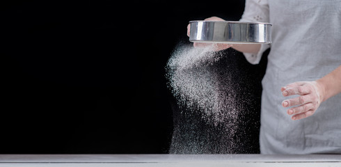Chef sifts flour through a sieve on a wooden table. Empty space for text. Isolated on dark background