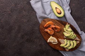 Wholegrain toast bread slices with guacamole, fried shrimp and fried bacon pieces on wooden board Selective Focus, Focus on the front of the shrimp on the first bread