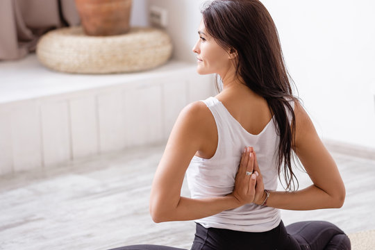 Rear view of an elegant young slim brunette woman doing baddha konasana sitting on a rug on the floor with her eyes closed in cozy quiet environment. Health and Body Improvement Concept. Home fitness
