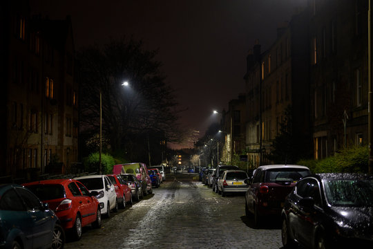 British urban streets at night. Dark alley of Edinburgh city with cars parking on the side of the road, damp from the rain. Downtown apartment buildings and homes neighborhood, street lights on.