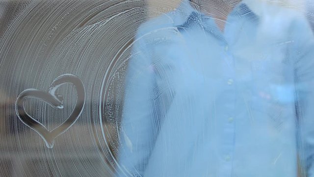 Woman cleaning window with detergent and rag drawing heart on soapy surface, fun