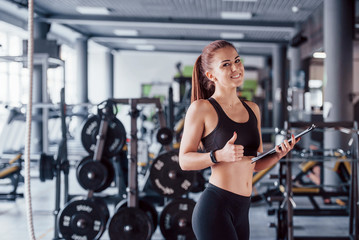 Young female fitness personal trainer with notepad standing in the gym with thumb up